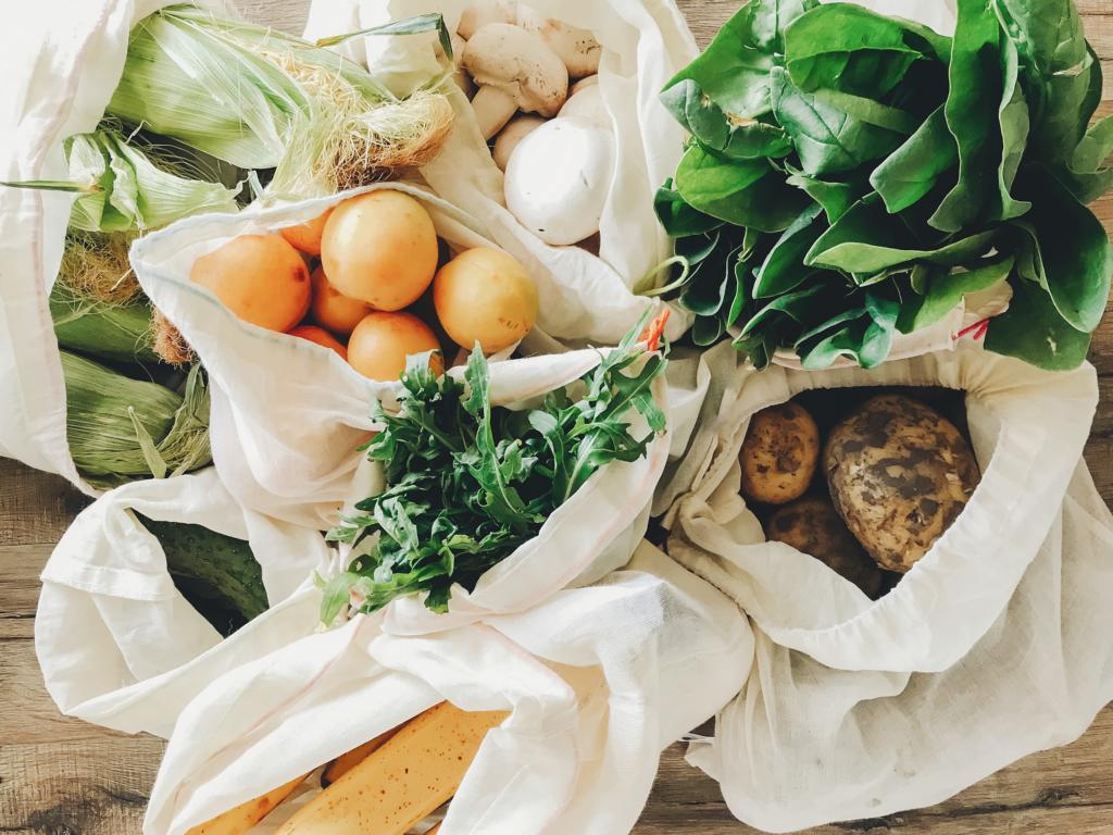 Tüten mit frischem Gemüse, darunter Mais, Pilze, Blattgemüse, Kartoffeln und verschiedene andere Produkte auf einer Holzoberfläche.