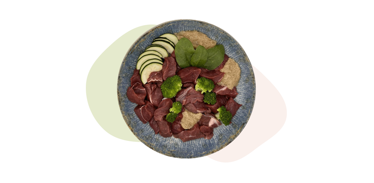 Eine blaue Schüssel mit Muskelfleisch vom Wild (Hirsch & Reh), Brokkoli, Spinatblättern, Zucchini.
