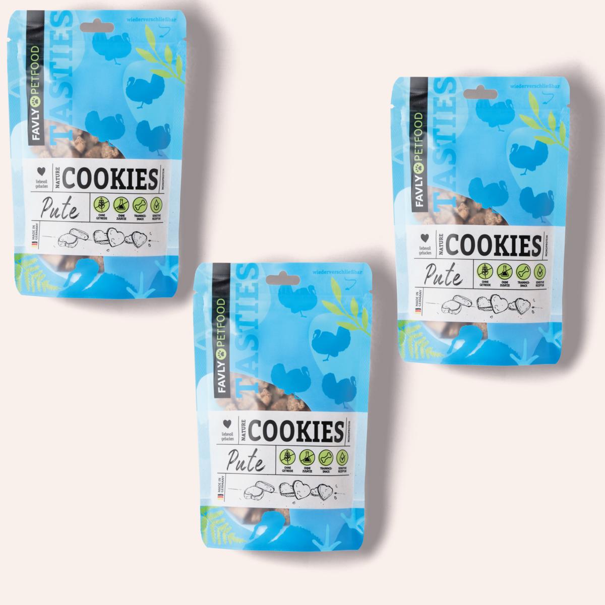 Drei Päckchen FAVLY Petfood Nature Cookies Pute für Hunde, in blau-weißer Verpackung, liegen auf einem hellbeigen Hintergrund.