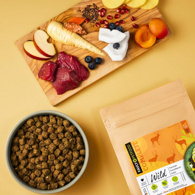 Eine Schüssel Trockenfutter Wild mit Pastinake, Preiselbeeren & Kokos von FAVLY Petfood, eine Packung Tierfutter und ein Schneidebrett mit verschiedenen Früchten, Gemüsesorten und Fleisch stehen auf einer gelben Oberfläche. Perfekt für ernährungssensible Hunde, die eine ausgewogene Mahlzeit suchen.