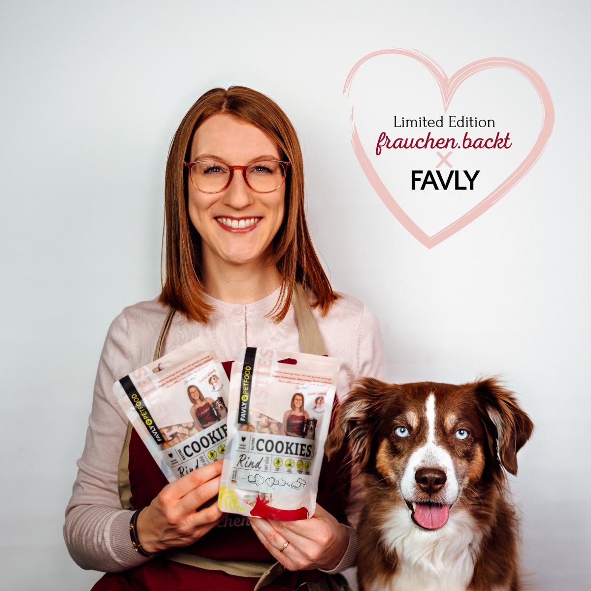 Eine Frau hält Päckchen mit „Limited Edition – Nature Cookies @frauchen.backt“ von FAVLY Petfood neben ihrem braun-weißen Hund, der gespannt auf seine Trainingsleckerlis wartet.