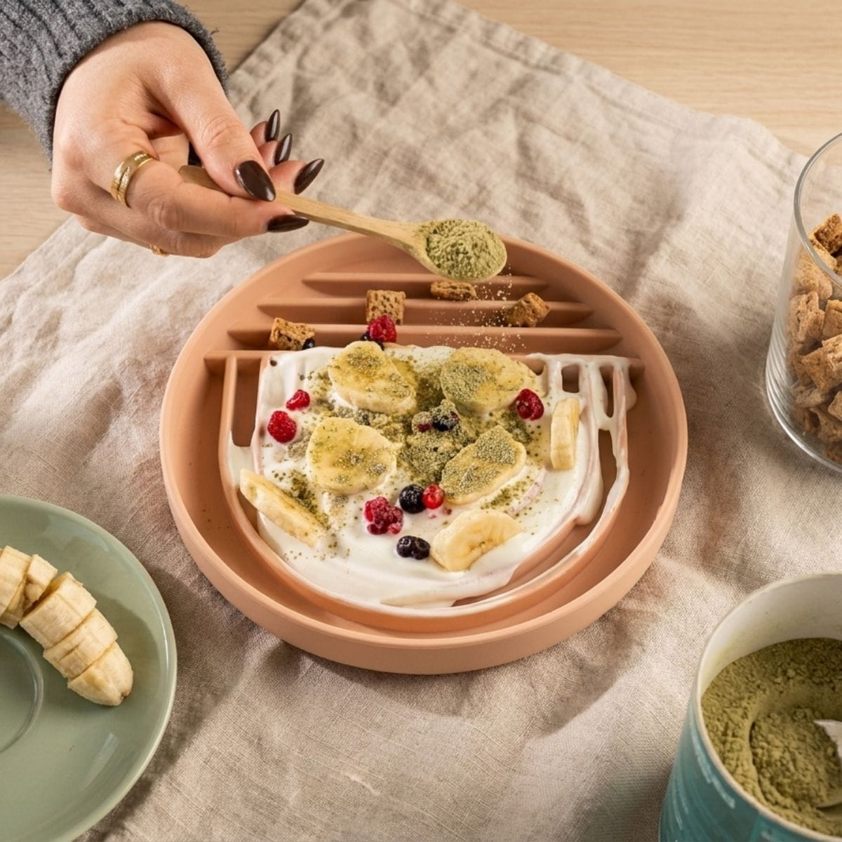 Eine Hand streut grünes Pulver über Joghurt und Obst auf einer FAVLY Petfood Pawfect Lick Mat – Schleckmatte.