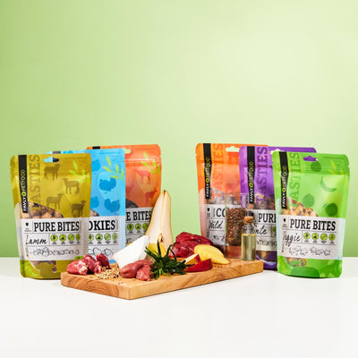Vier FAVLY Probierpaket "Snack it" Tüten von FAVLY Petfood auf Holzbrett vor grünem Hintergrund.
